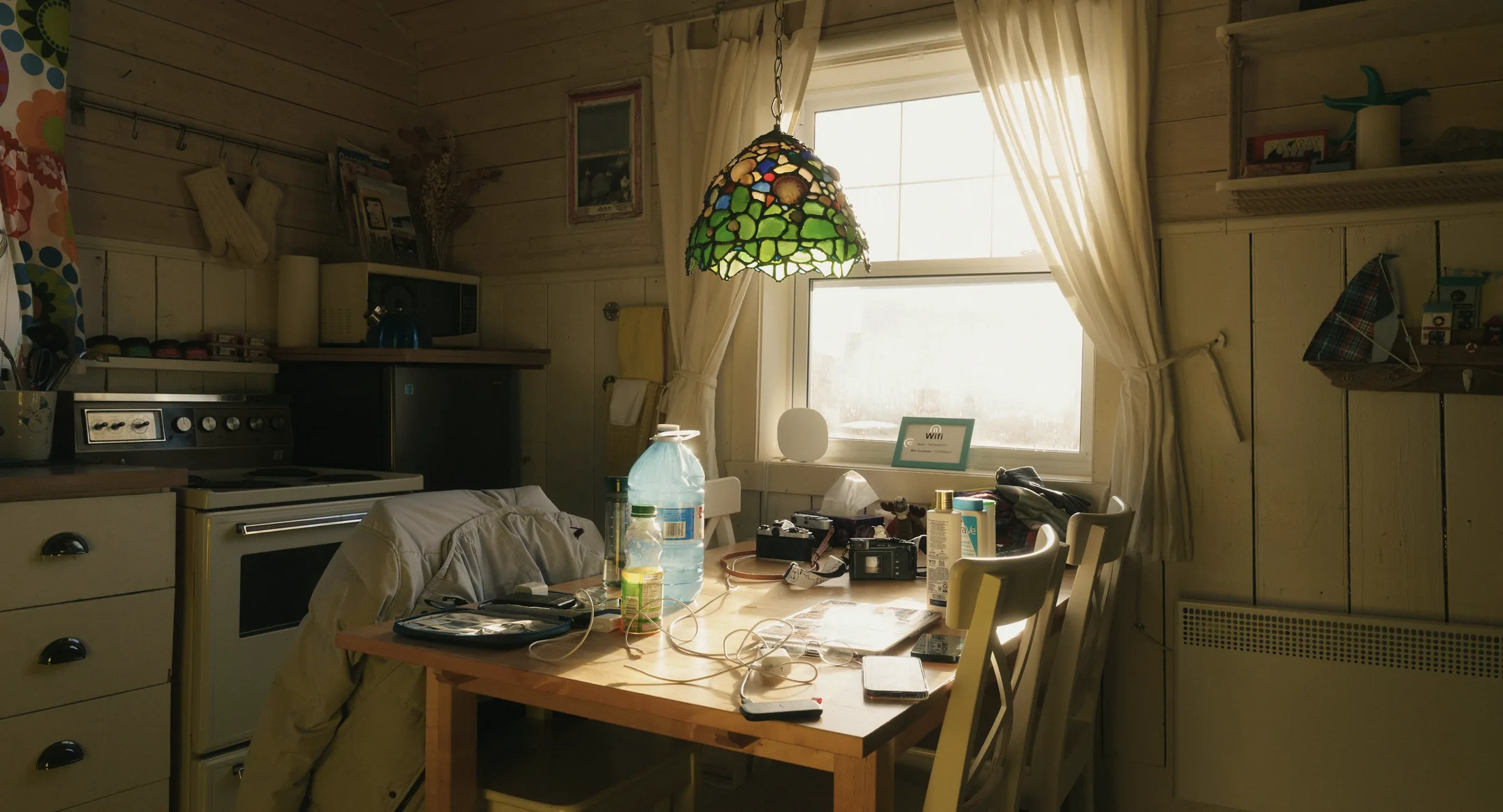 A sunlit, lived-in kitchen table holding cameras, papers, cables, and everyday belongings.